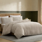 A beige and cream striped bedding set with a textured basketweave pattern, styled on a bed in a modern bedroom with a dark green wainscoted wall and a rattan headboard.