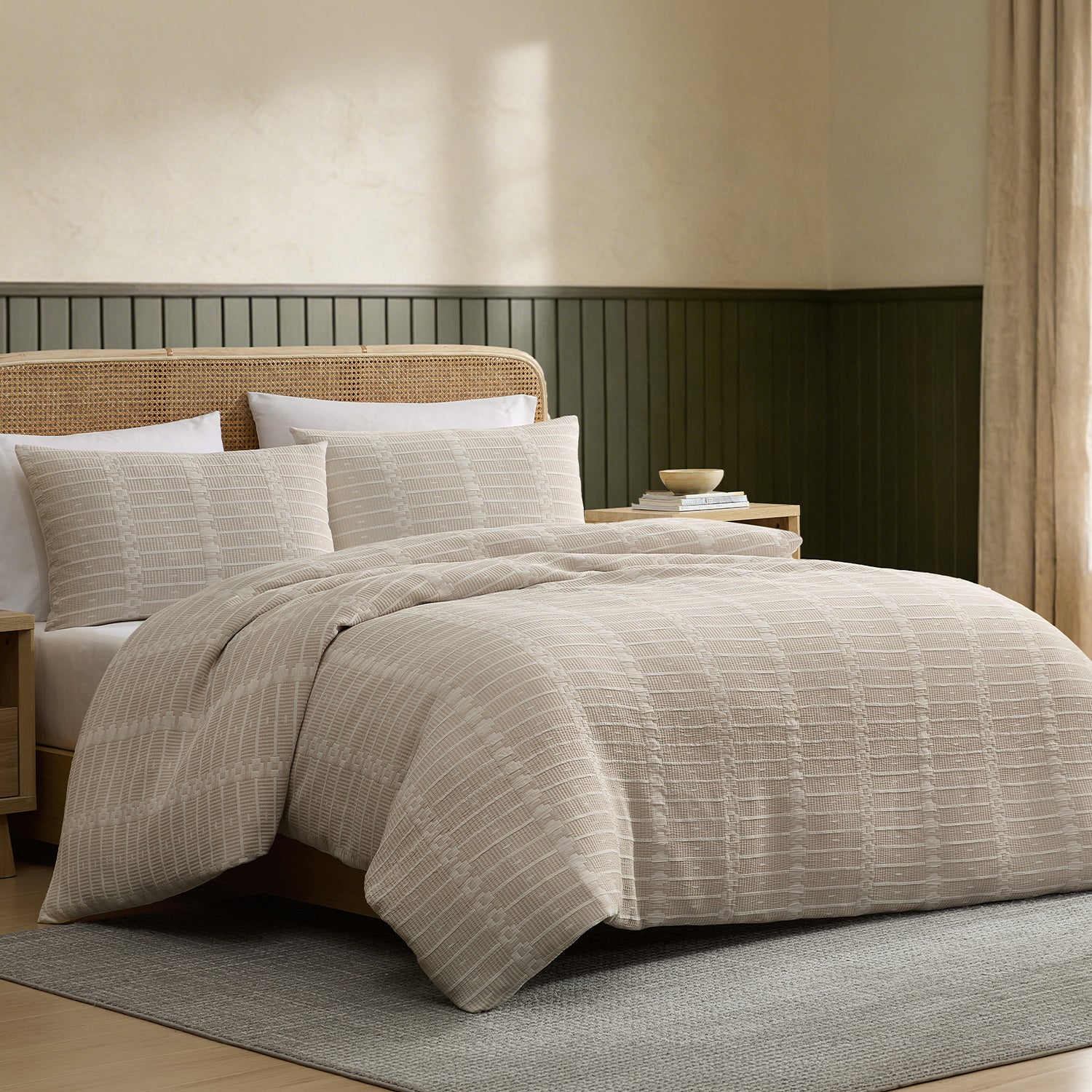 A beige and cream striped bedding set with a textured basketweave pattern, styled on a bed in a modern bedroom with a dark green wainscoted wall and a rattan headboard.