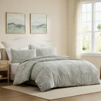 A light sage green quilted comforter set with matching pillow shams on a bed in a softly lit beige bedroom. Two framed landscape prints hang above the headboard, and a window with sheer curtains is visible on the right.
