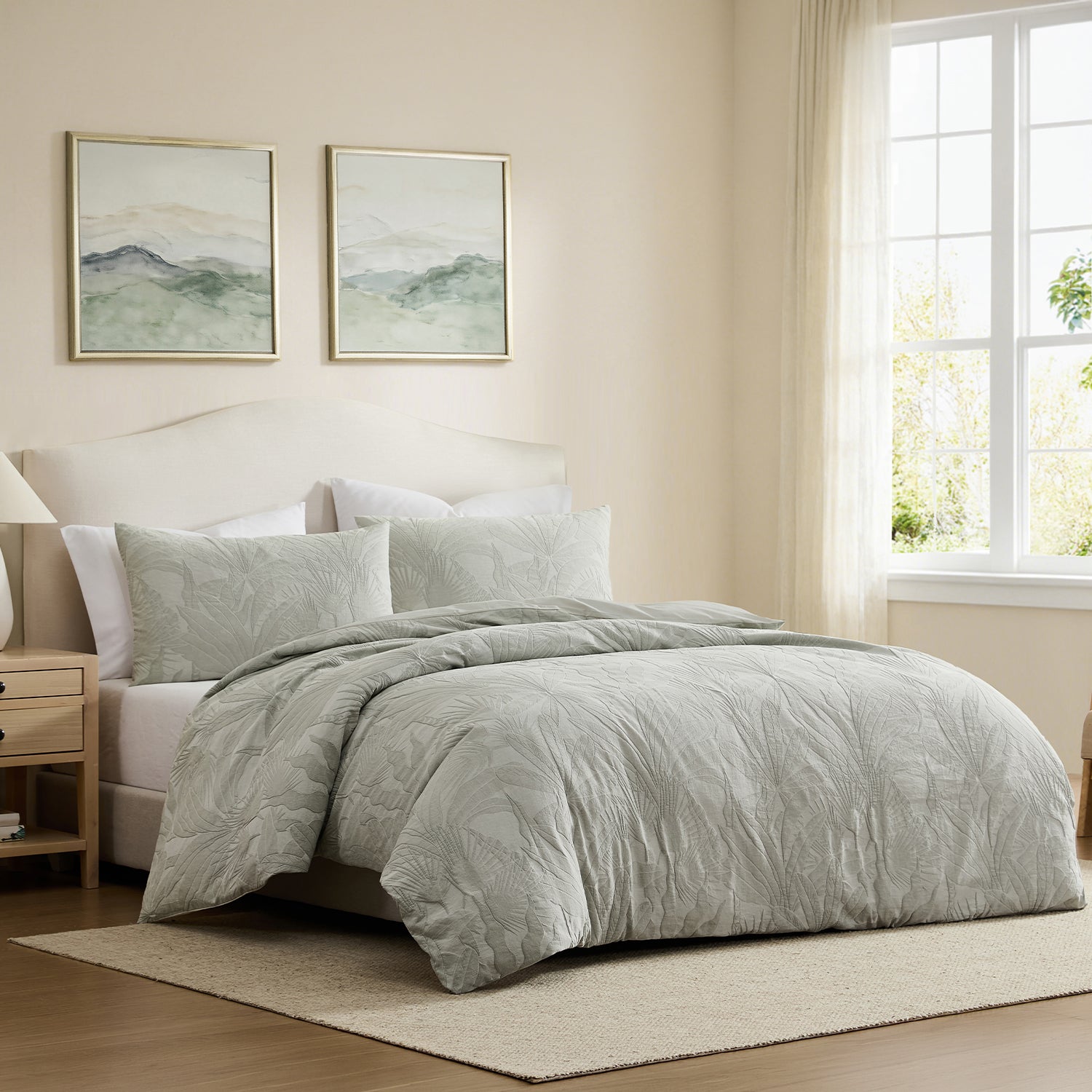 A light sage green quilted comforter set with matching pillow shams on a bed in a softly lit beige bedroom. Two framed landscape prints hang above the headboard, and a window with sheer curtains is visible on the right.