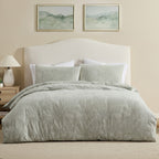 A neatly made bed with a light sage green patterned comforter and matching pillow shams, set against a beige upholstered headboard in a softly lit bedroom. Two framed landscape prints hang above the bed, and a bedside lamp sits on the left.