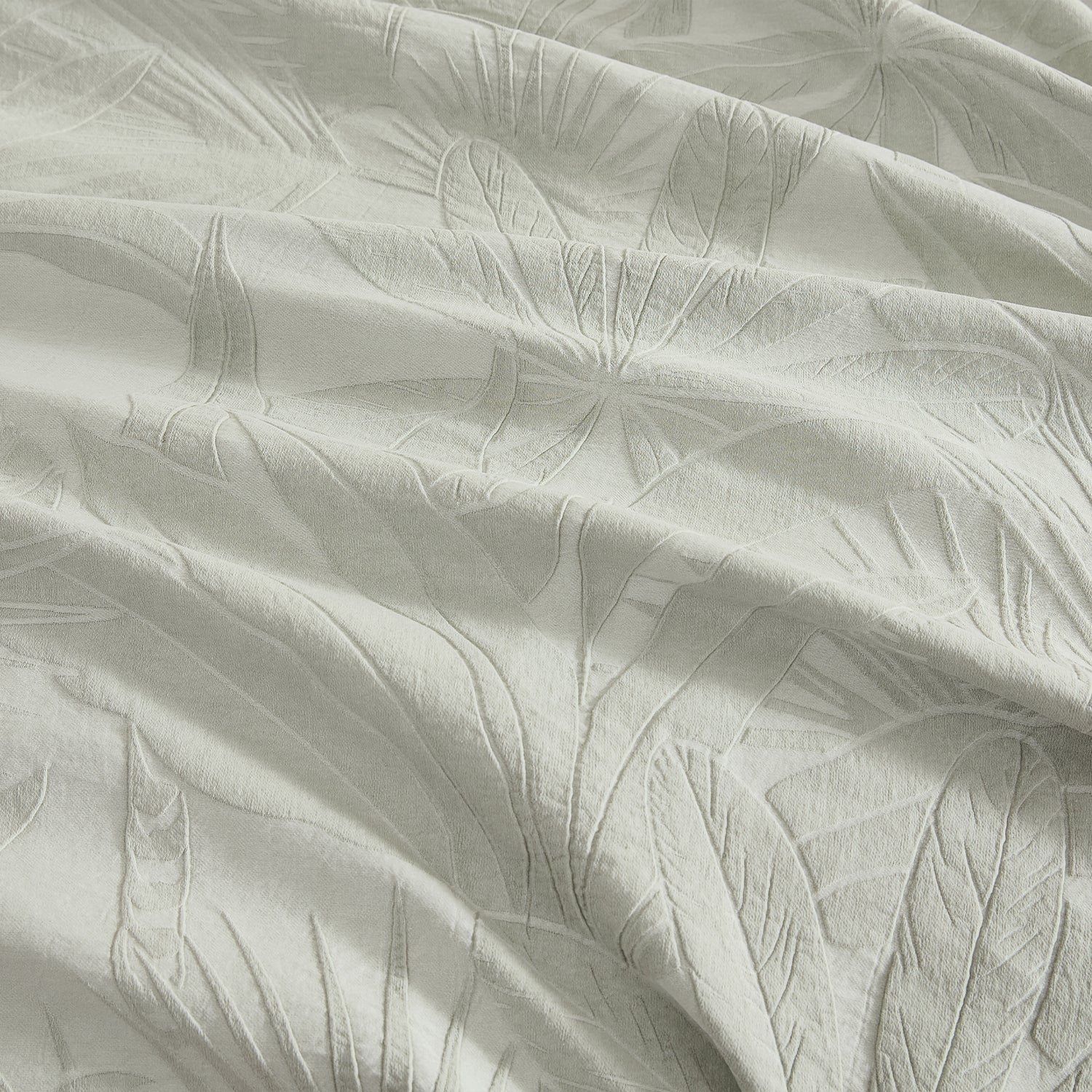 A close-up of textured fabric with a tonal leaf pattern, shown in soft folds with a light gray-green hue.