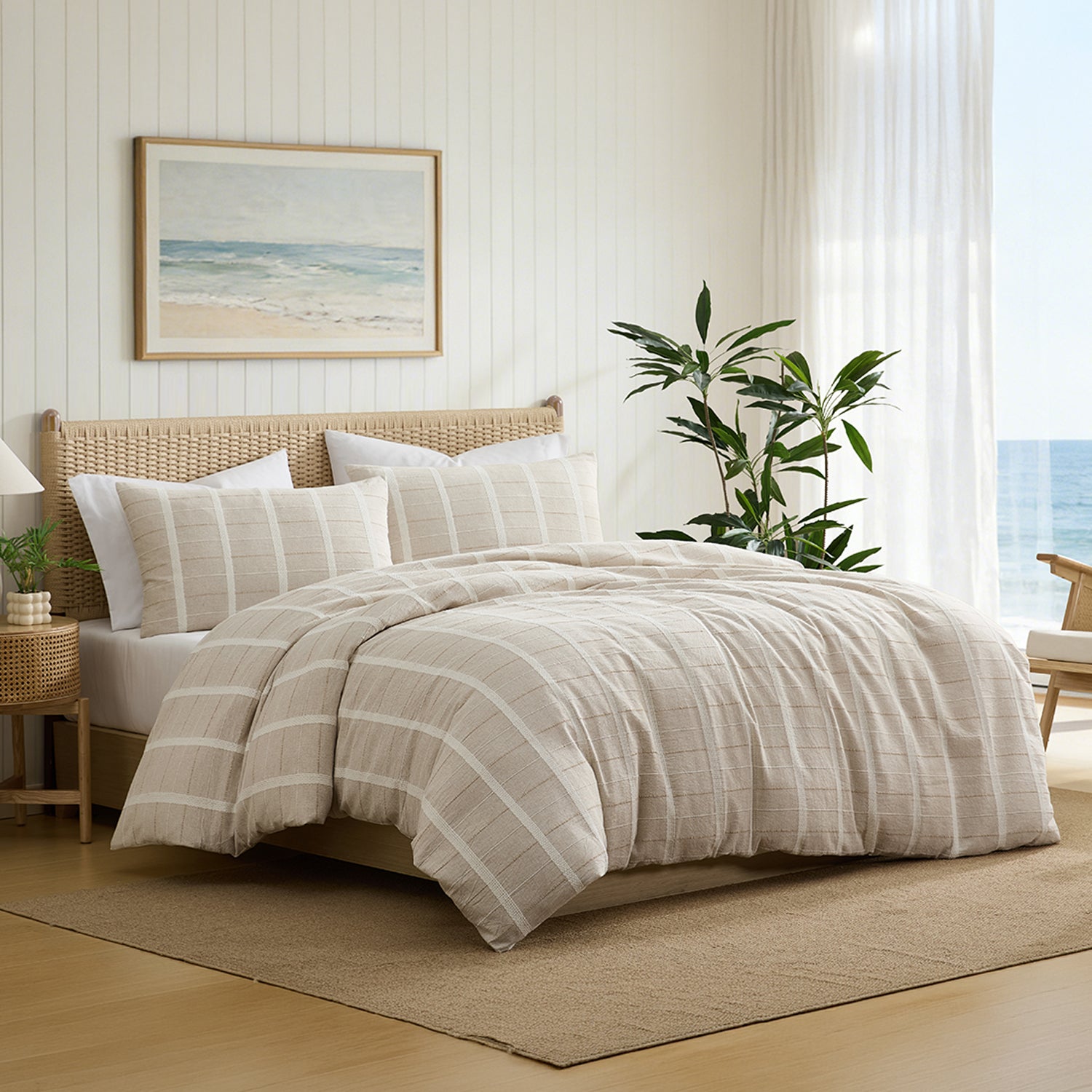 A beige and white striped bedding set with matching shams is styled on a bed in a bright, modern beach-themed bedroom. The room features white paneled walls, a framed beach print, a potted green plant, and a view of the ocean.