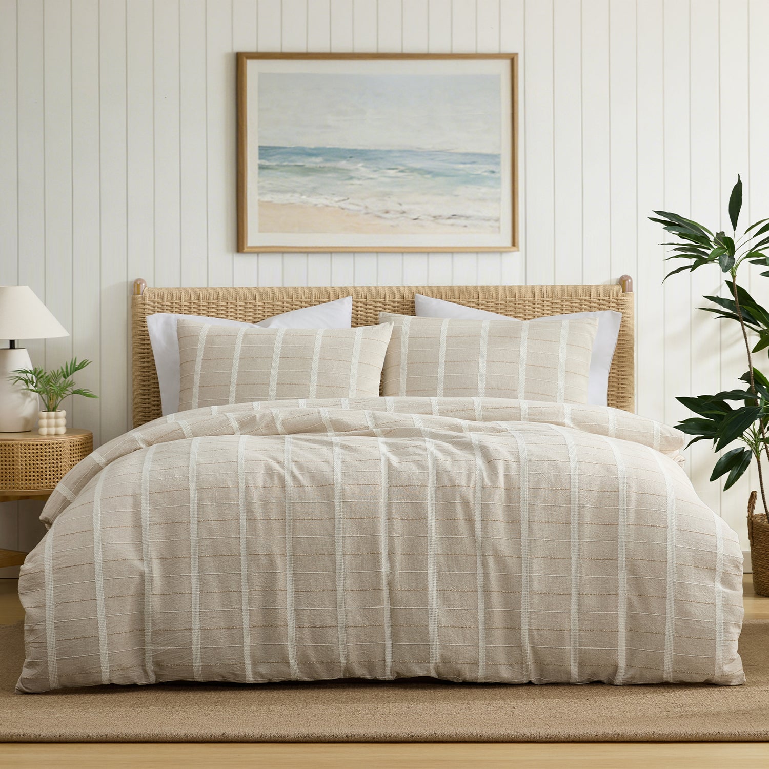 A neatly made bed with a beige and white vertically striped duvet cover and matching pillow shams, styled in a bright bedroom with a wicker headboard, wall art, bedside lamp, and houseplant.