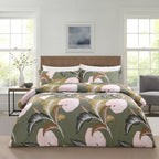 A bed dressed in a tropical floral bedding set with a sage green background, large pink flowers, and gray-green leaves, styled in a bright bedroom with two windows, a wooden headboard, and a side table.