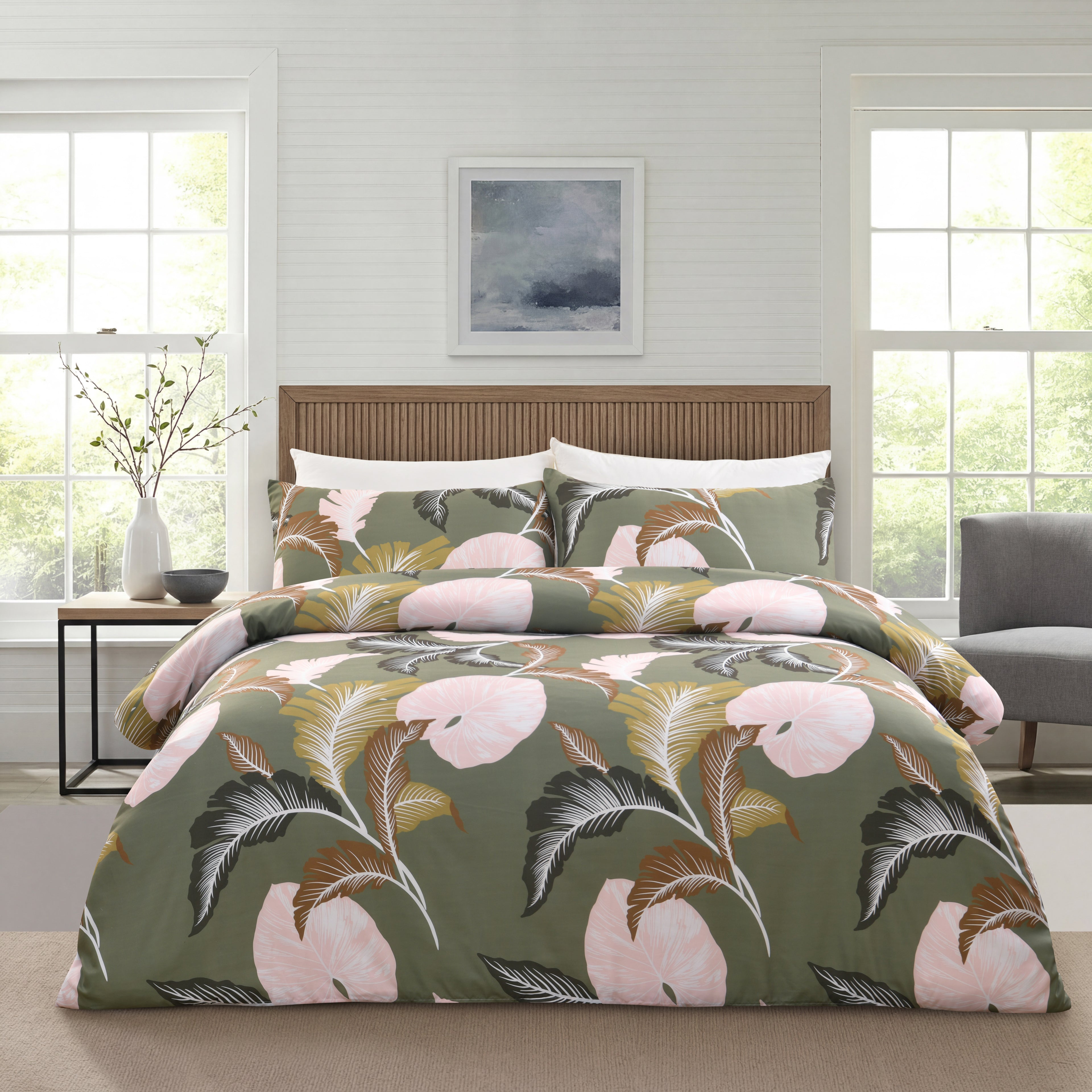 A bed dressed in a tropical floral bedding set with a sage green background, large pink flowers, and gray-green leaves, styled in a bright bedroom with two windows, a wooden headboard, and a side table.