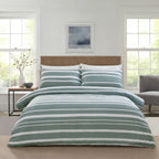 A neatly made bed in a bright bedroom setting, featuring a striped duvet cover and matching pillow shams in sage green and white. The room has white paneled walls, a wooden slat headboard, a small side table with a vase, and a gray armchair.