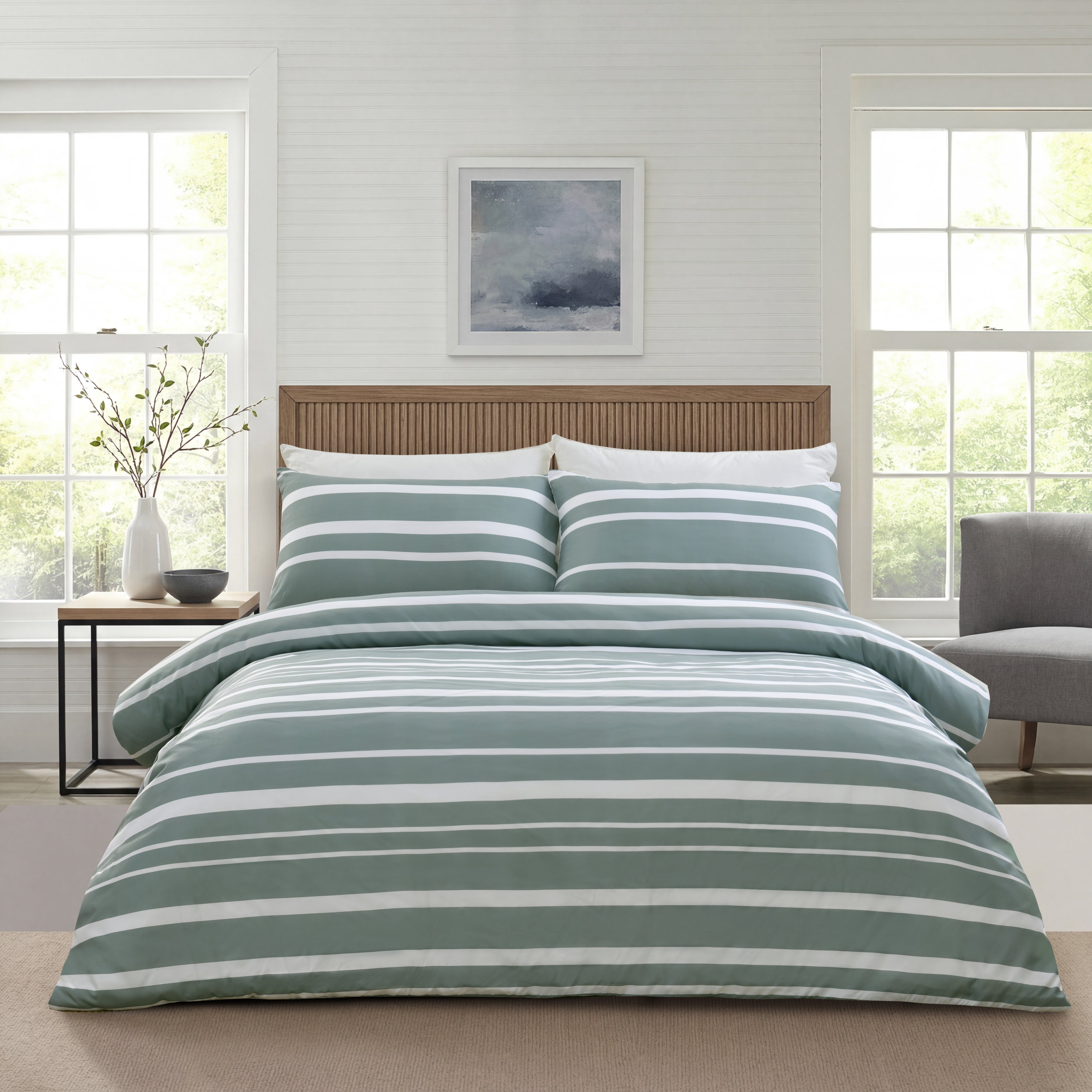 A neatly made bed in a bright bedroom setting, featuring a striped duvet cover and matching pillow shams in sage green and white. The room has white paneled walls, a wooden slat headboard, a small side table with a vase, and a gray armchair.