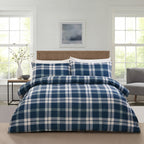 A bed dressed in a navy blue and white plaid duvet cover and matching pillow shams, styled in a bright, modern bedroom with white paneled walls, two windows, and a wooden headboard.