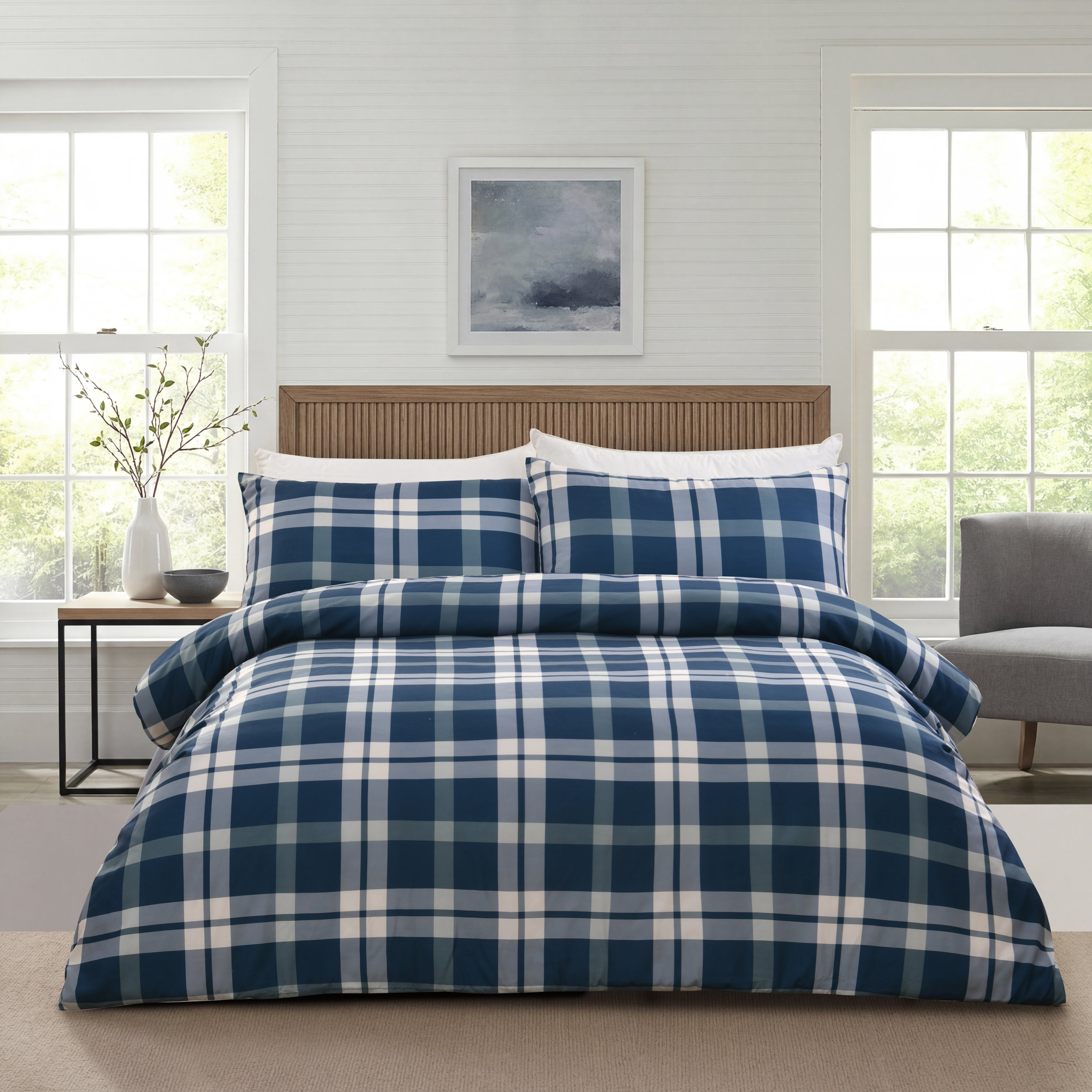 A bed dressed in a navy blue and white plaid duvet cover and matching pillow shams, styled in a bright, modern bedroom with white paneled walls, two windows, and a wooden headboard.