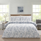 A neatly made bed in a bright bedroom setting, featuring a white duvet cover and matching pillow shams with a delicate blue-gray botanical floral pattern. The bed is styled with a light wood headboard, flanked by a small side table with a vase and a gray armchair in the background.