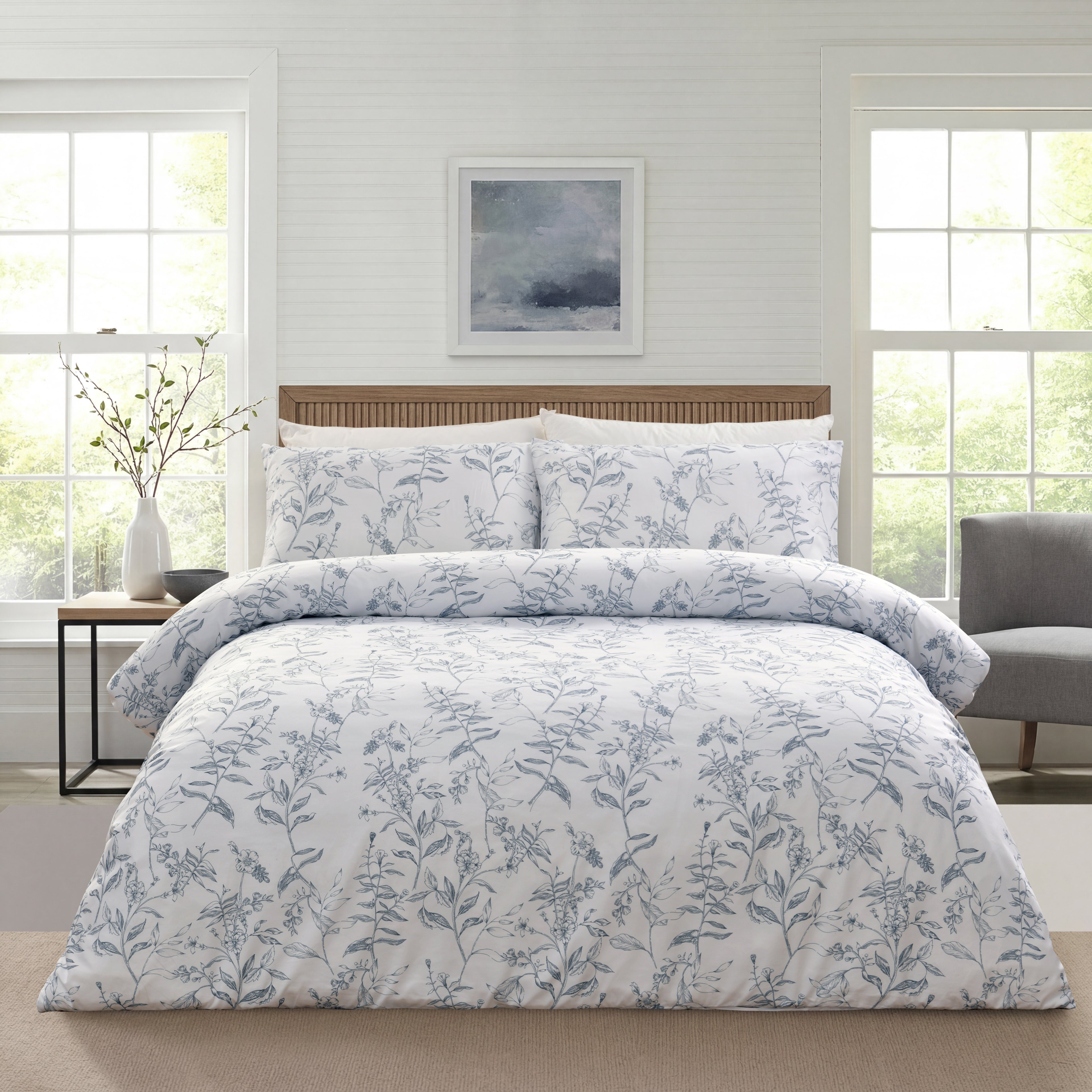 A neatly made bed in a bright bedroom setting, featuring a white duvet cover and matching pillow shams with a delicate blue-gray botanical floral pattern. The bed is styled with a light wood headboard, flanked by a small side table with a vase and a gray armchair in the background.