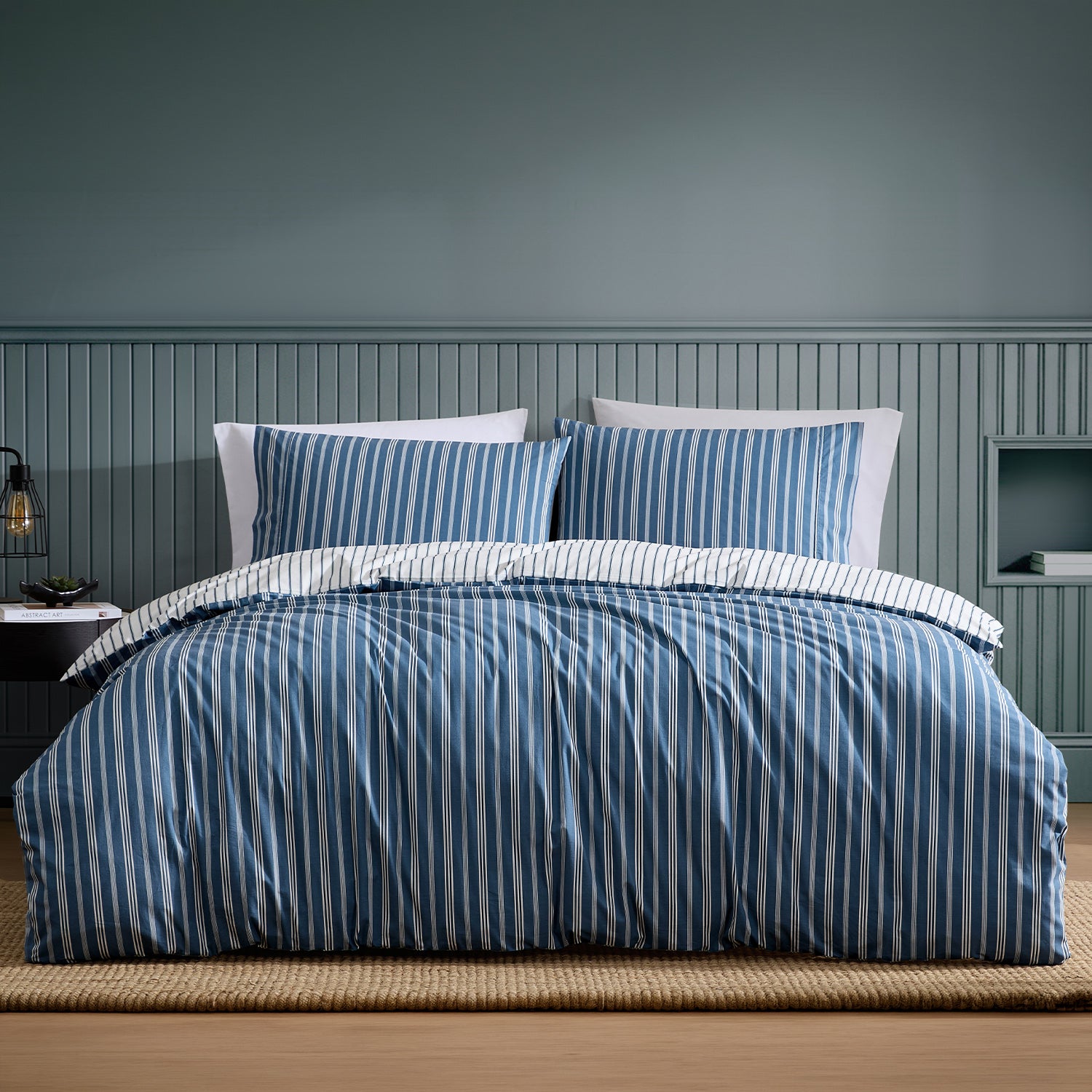 A neatly made bed with a blue and white striped duvet cover and matching pillow shams, set against a blue paneled wall in a styled bedroom.