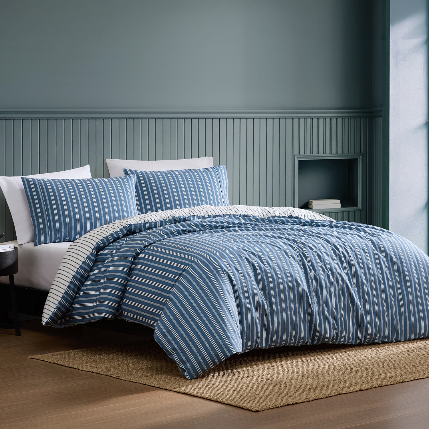 A bed dressed in a blue and white striped duvet cover and matching pillow shams, styled in a bedroom with blue paneled walls, wood flooring, and a woven rug.
