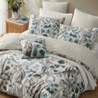 A made bed with a coordinated bedding set featuring a botanical leaf print in muted green, gray, and blue-gray tones on a light background, with matching pillow shams and a small accent pillow. The room includes a woven side table with flowers and a small blue vase against a warm beige wall.