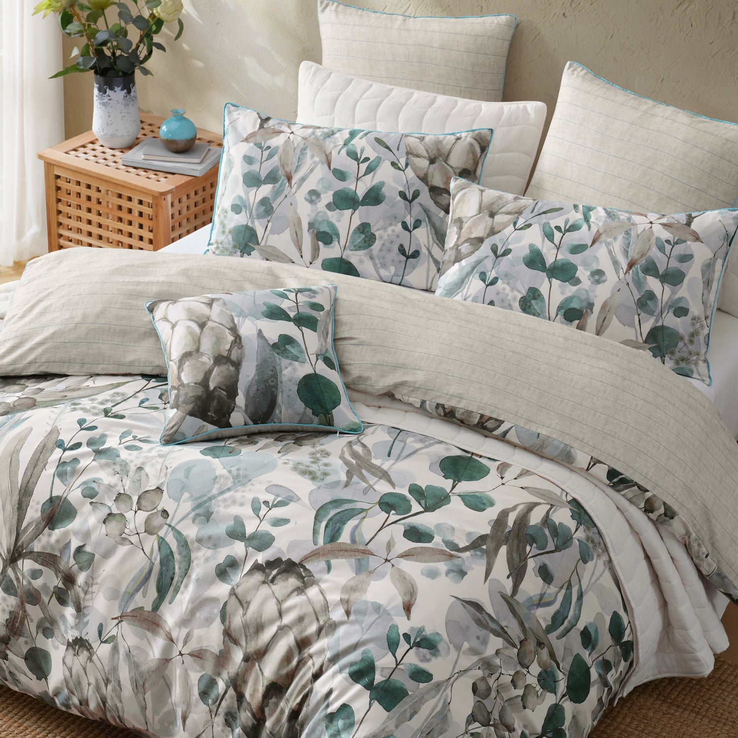 A made bed with a coordinated bedding set featuring a botanical leaf print in muted green, gray, and blue-gray tones on a light background, with matching pillow shams and a small accent pillow. The room includes a woven side table with flowers and a small blue vase against a warm beige wall.