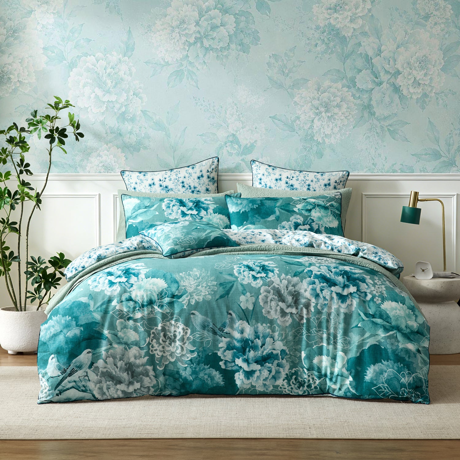 A soft blue teal and white floral bedding set styled on a bed in a bedroom with matching floral wallpaper, white wainscoting, a potted plant, and a bedside lamp.