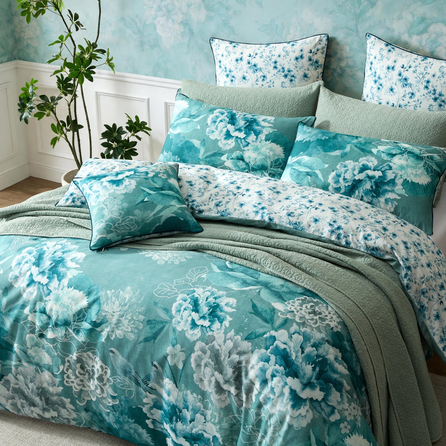 A floral bedding set styled on a bed, featuring a duvet cover and matching pillowcases in shades of teal, blue, and white with large peony-like flowers, accented by solid sage green pillows and a matching sage green throw blanket.