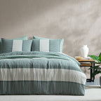 A bed dressed in a striped bedding set with teal, white, and gray vertical stripes, styled in a bedroom setting with a side table, white vase, and green plant against a textured beige wall.