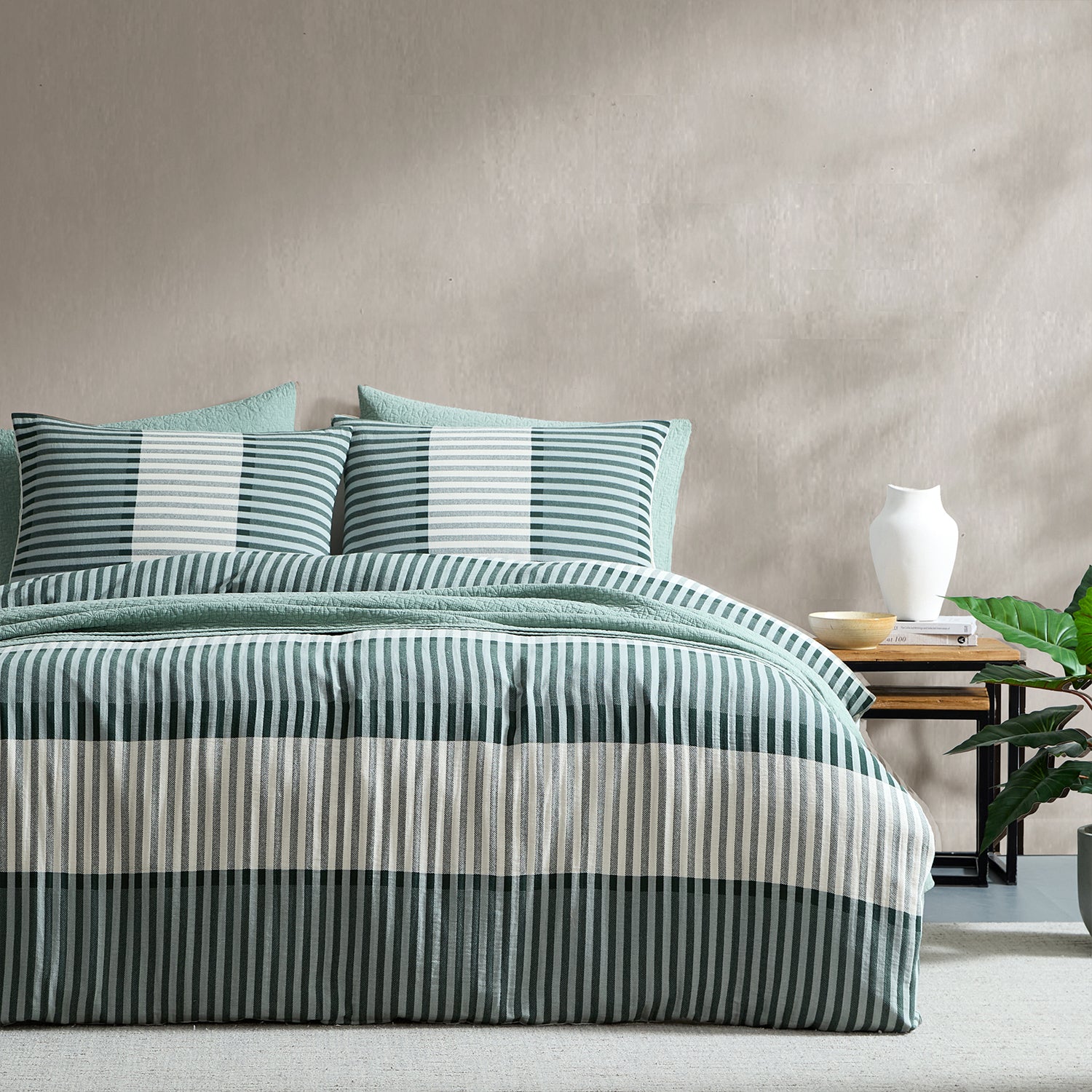 A bed dressed in a striped bedding set with teal, white, and gray vertical stripes, styled in a bedroom setting with a side table, white vase, and green plant against a textured beige wall.