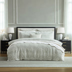 A neatly made bed in a styled bedroom, featuring a light gray and white patterned quilt, matching shams, and white pillows. The bed is flanked by matching nightstands with white table lamps, set against a gray paneled wall.