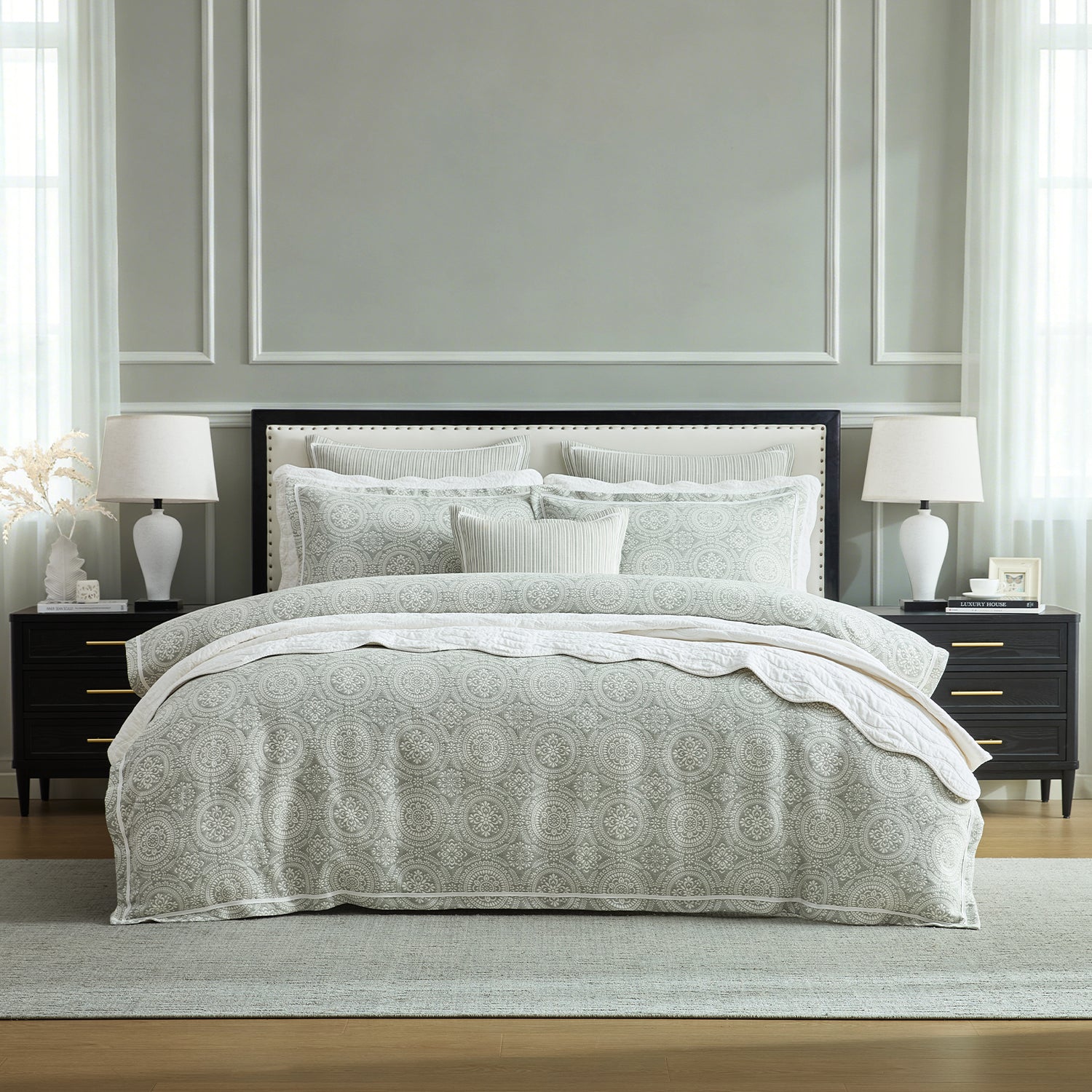 A neatly made bed in a styled bedroom, featuring a light gray and white patterned quilt, matching shams, and white pillows. The bed is flanked by matching nightstands with white table lamps, set against a gray paneled wall.
