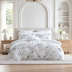 A neatly made bed in a bright, elegant bedroom featuring a white and gray floral print bedding set with multiple pillows and a duvet cover.