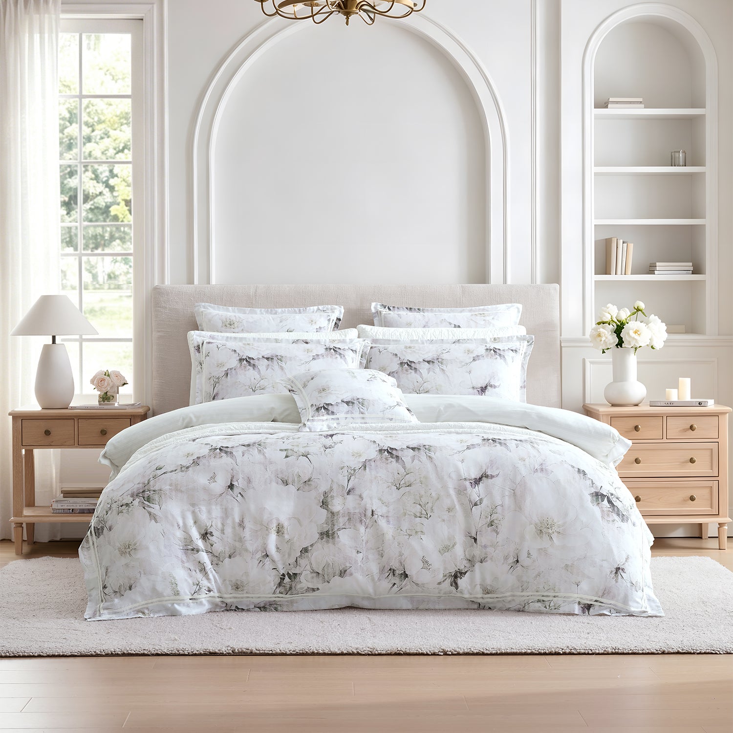 A neatly made bed in a bright, elegant bedroom featuring a white and gray floral print bedding set with multiple pillows and a duvet cover.
