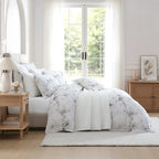 A neatly made bed in a bright bedroom featuring a white bedding set with a delicate gray floral print. The room includes light wood furniture, sheer white curtains, and a large window looking out onto green trees.