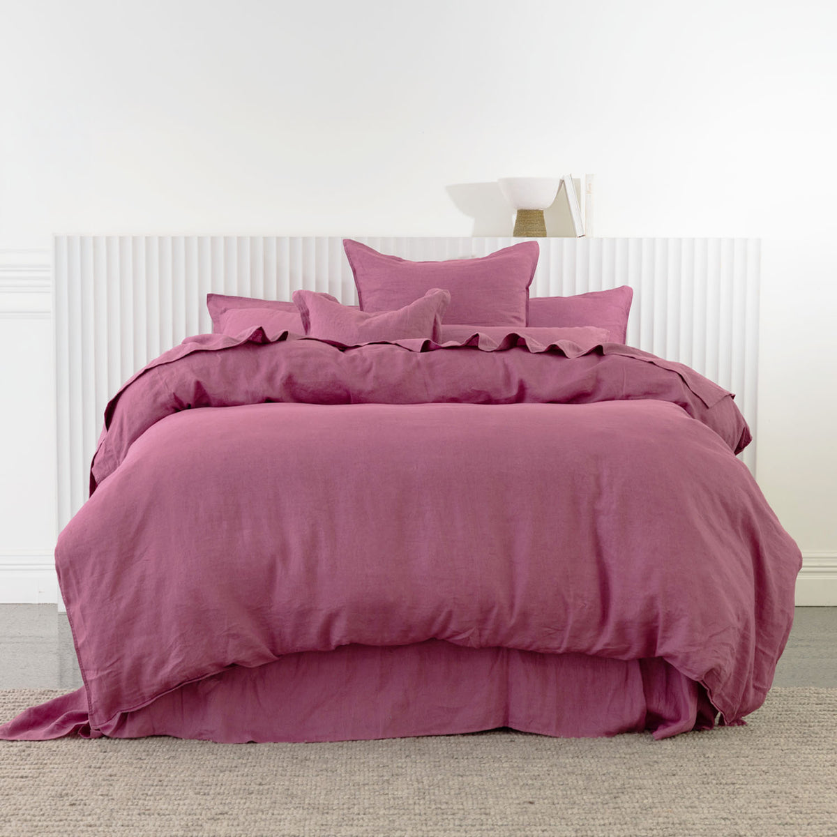 A bed dressed in a berry bedding set with matching pillows and a ruffled duvet cover, set against a white paneled headboard and light gray carpet.