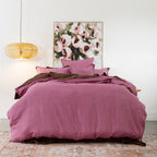 A bed dressed in a berry hue bedding set with matching pillows and a dark brown under-sheet, styled in a bedroom setting with a wicker pendant light, a floral painting, and a patterned rug.