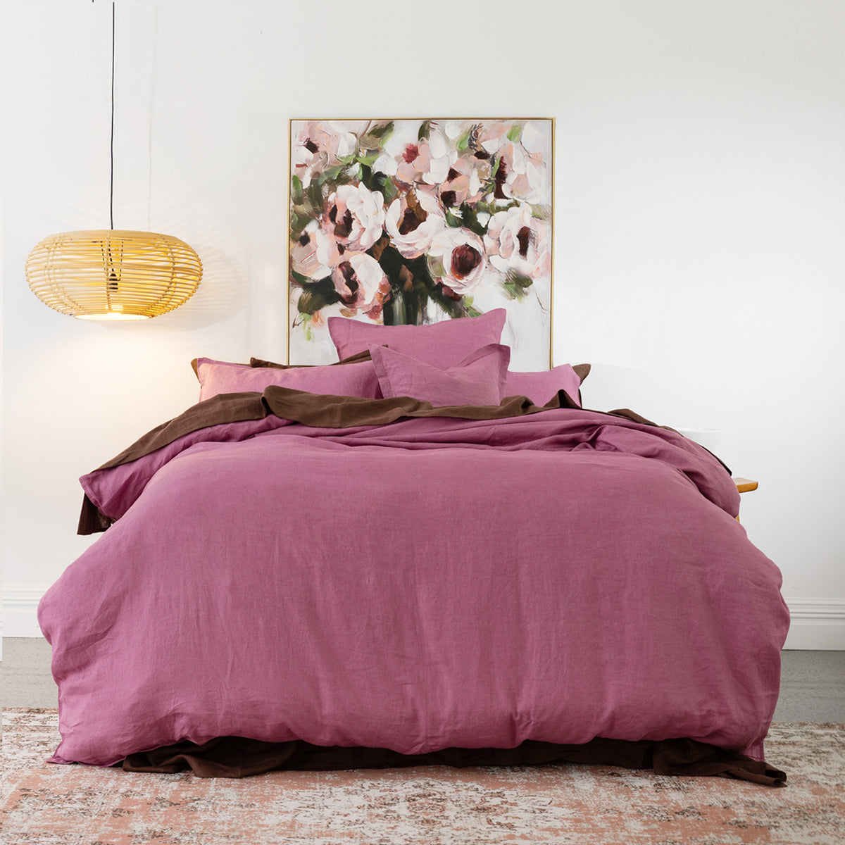 A bed dressed in a berry hue bedding set with matching pillows and a dark brown under-sheet, styled in a bedroom setting with a wicker pendant light, a floral painting, and a patterned rug.