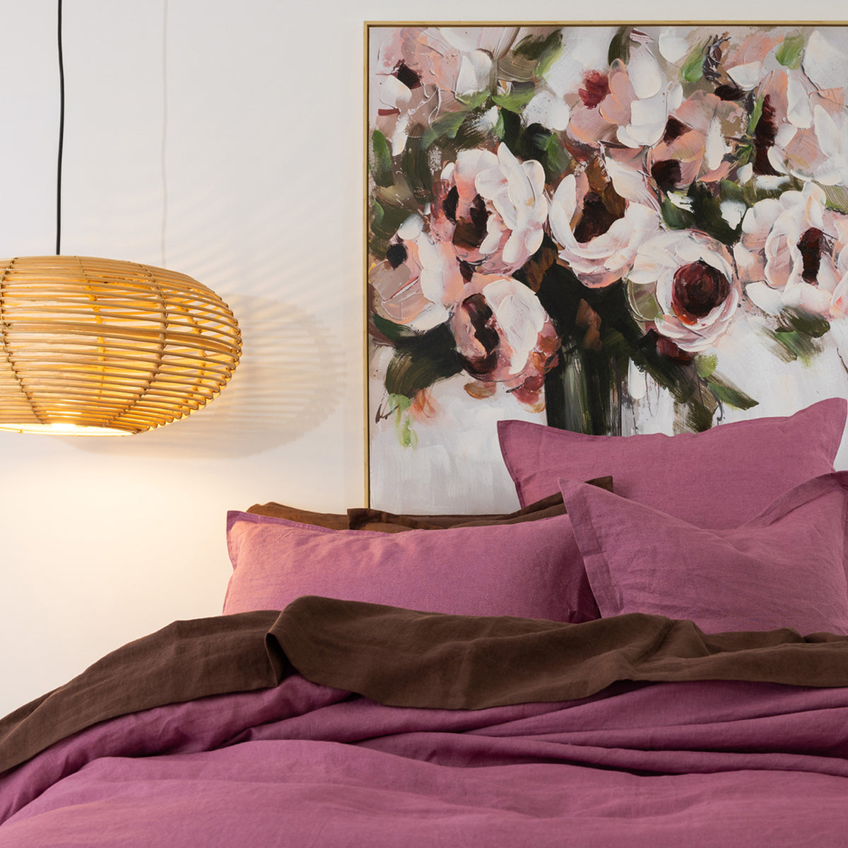 A styled bedroom scene with a bed dressed in berry bedding and pillows, with a brown sheet draped across the foot. A large framed floral painting hangs on the wall above the bed, and a woven pendant lamp glows on the left against a white wall.