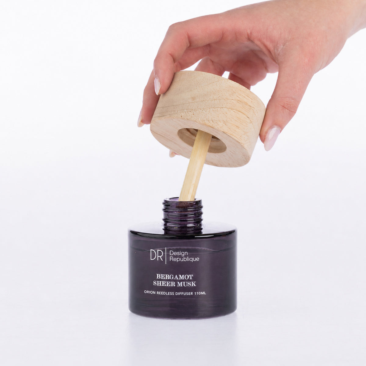 Design Republique Bergamot Sheer Musk Orion Reedless Diffuser shown open with the natural wood-look lid and bamboo diffuser insert being lifted from the purple glass vessel.