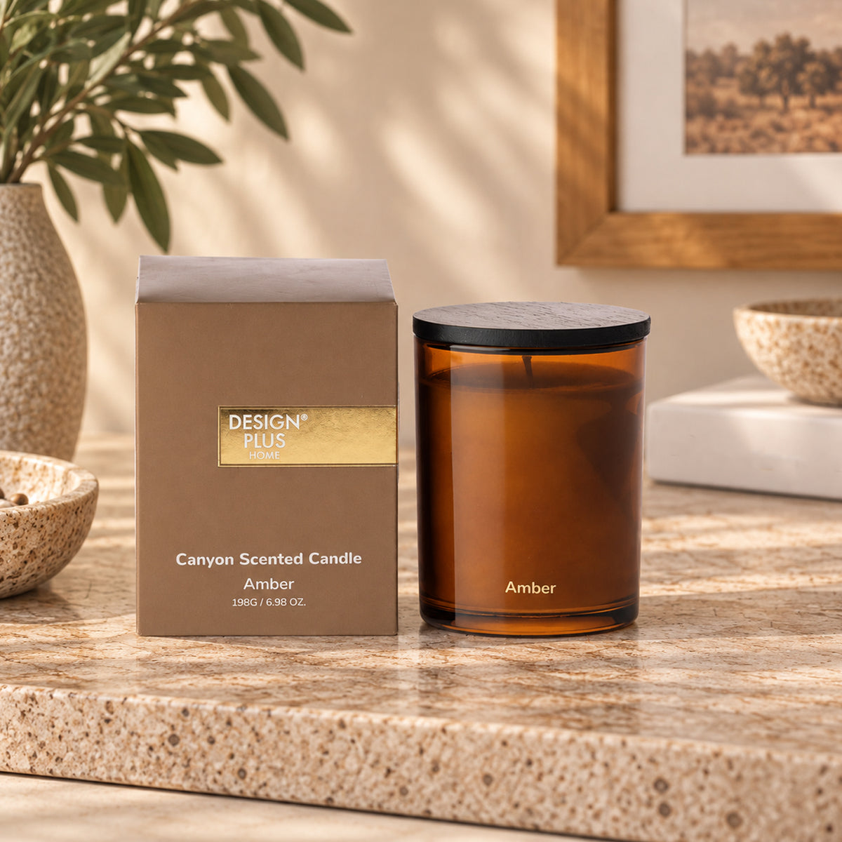 An amber scented candle in a glass jar with a black lid sits on a stone countertop beside its box. The box reads 'DESIGN PLUS CANDLE' and 'Canyon Scented Candle Amber'.
