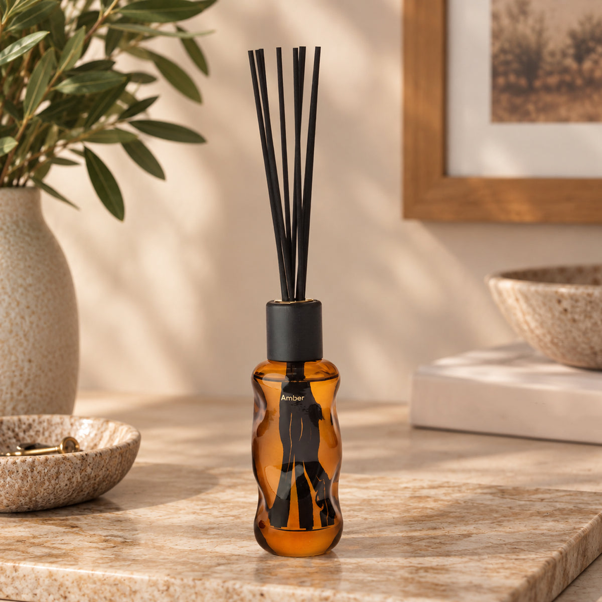 An amber glass reed diffuser bottle with a black cap and black reeds, placed on a stone countertop in a styled interior setting with a vase, bowl, framed art, and books in the background.