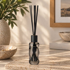 A reed diffuser bottle with black reeds and a dark smoky glass body sits on a stone surface in a styled interior setting with a plant, bowls, and framed art in the background.