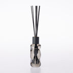 A reed diffuser bottle made of smoky translucent glass with a matte black cap and several black reeds, shown on a clean white background.