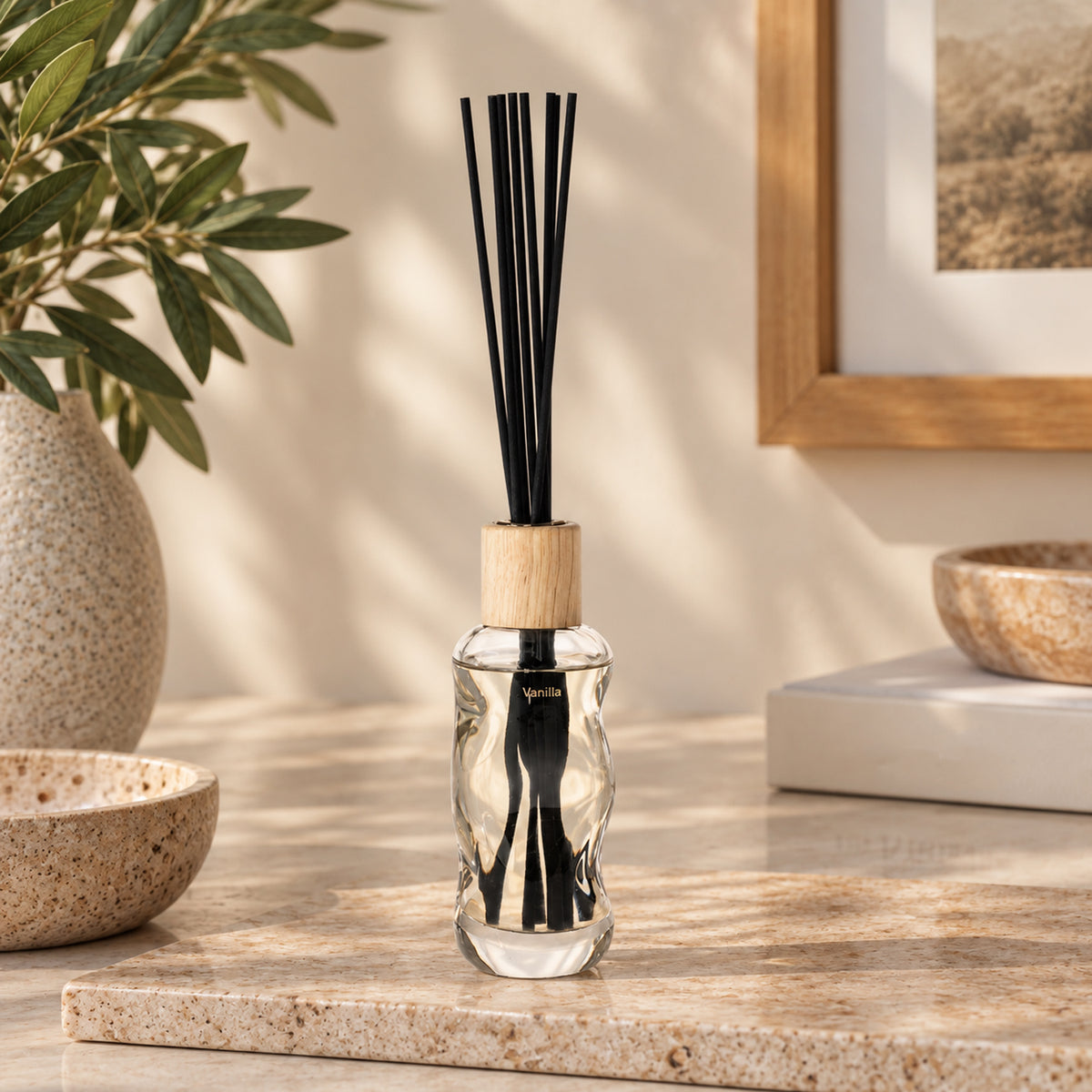 A clear glass reed diffuser bottle with a light wood cap and black reeds, styled on a stone surface with a plant, bowls, and framed art in a warm indoor setting.