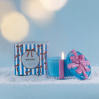 Christmas Wishes Bow Candle in a Box 110g Biscotti – Blue ribbed scented soy candle with a pink ribbon detail and decorative bow lid, displayed beside its coordinating blue and white striped Biscotti gift box amongst faux snow.