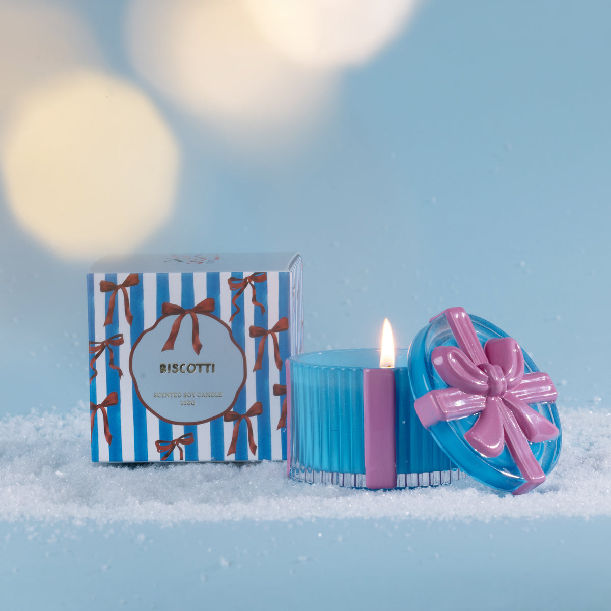 Christmas Wishes Bow Candle in a Box 110g Biscotti – Blue ribbed scented soy candle with a pink ribbon detail and decorative bow lid, displayed beside its coordinating blue and white striped Biscotti gift box amongst faux snow.