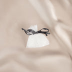 Eden Magnolia Scented Sachet 12g featuring a small white mesh pouch filled with fragrance beads and tied with a dark grey ribbon, displayed on beige satin fabric.