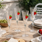 Four clear champagne flutes with raised strawberry embellishments displayed on a rustic outdoor table setting with fresh strawberries, grapes, cheese, and crackers.