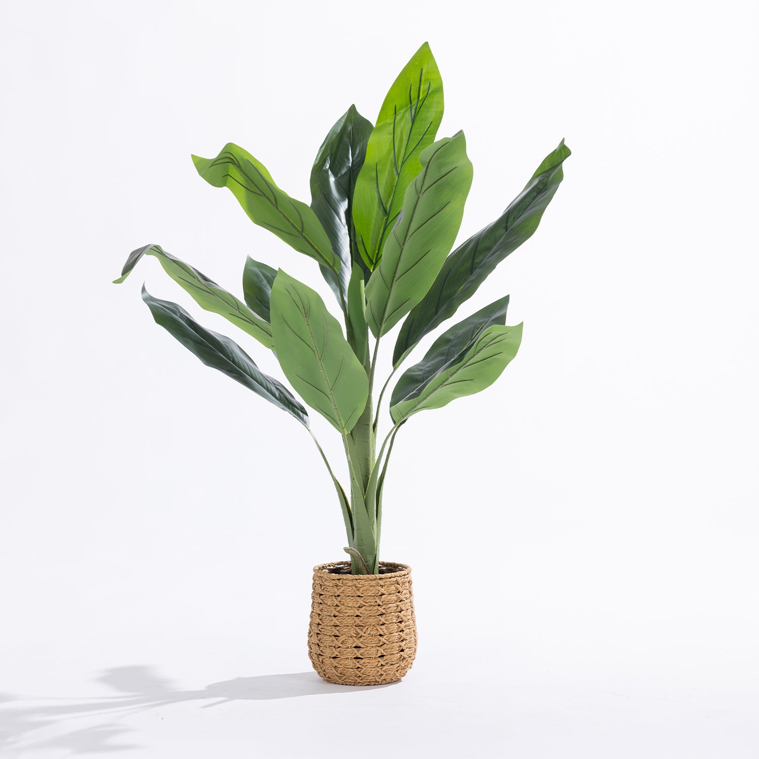 An artificial potted plant with large green leaves in a woven natural fiber basket pot, set against a plain white studio background.