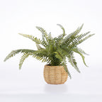 An artificial green fern plant in a round, light brown woven wicker basket pot, photographed against a seamless white studio background.