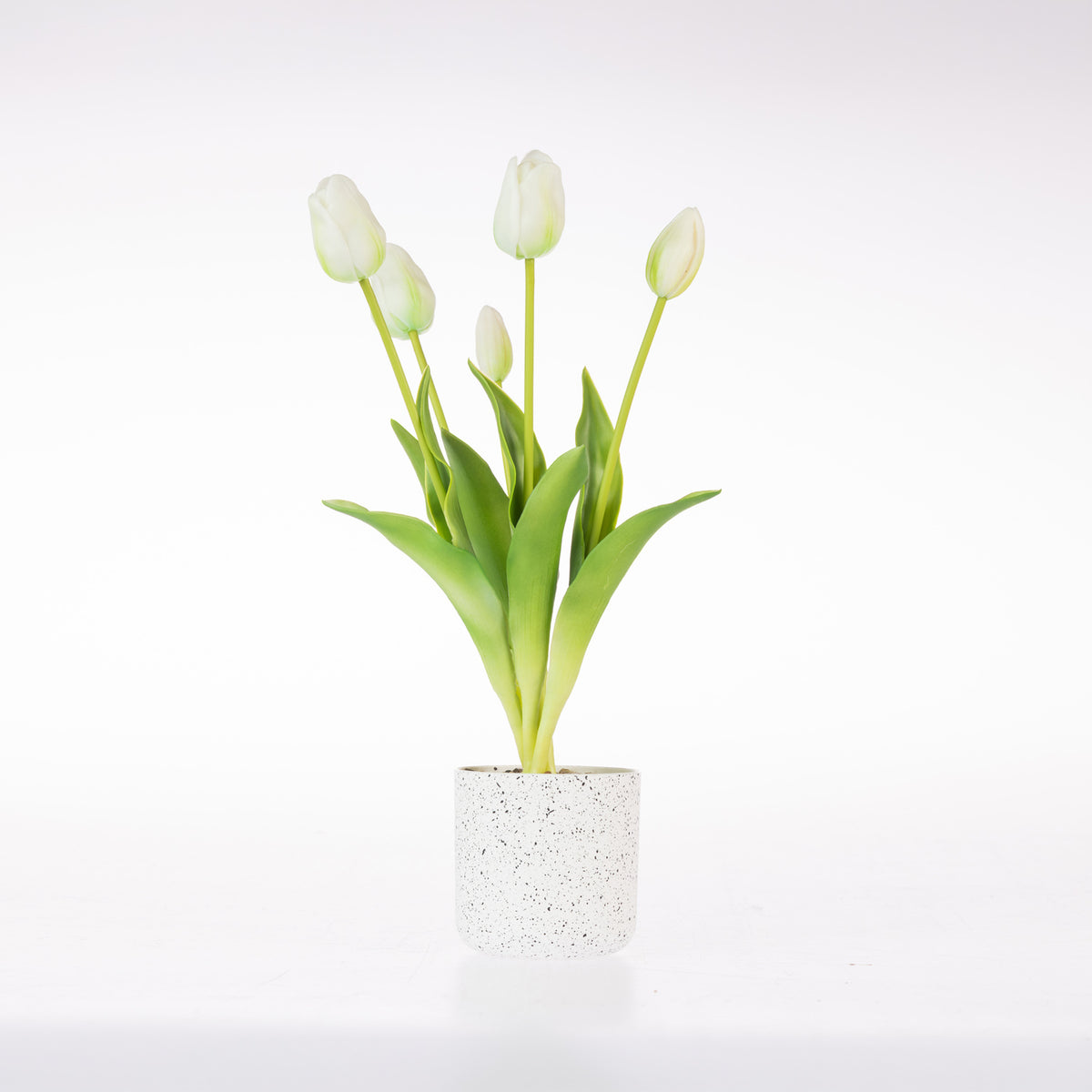 A small artificial tulip arrangement with five white tulip buds and green leaves in a speckled white cylindrical pot, photographed on a clean white background.