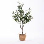 An artificial olive tree with green leaves and small dark berries, potted in a natural woven basket with side handles, photographed against a plain white background.