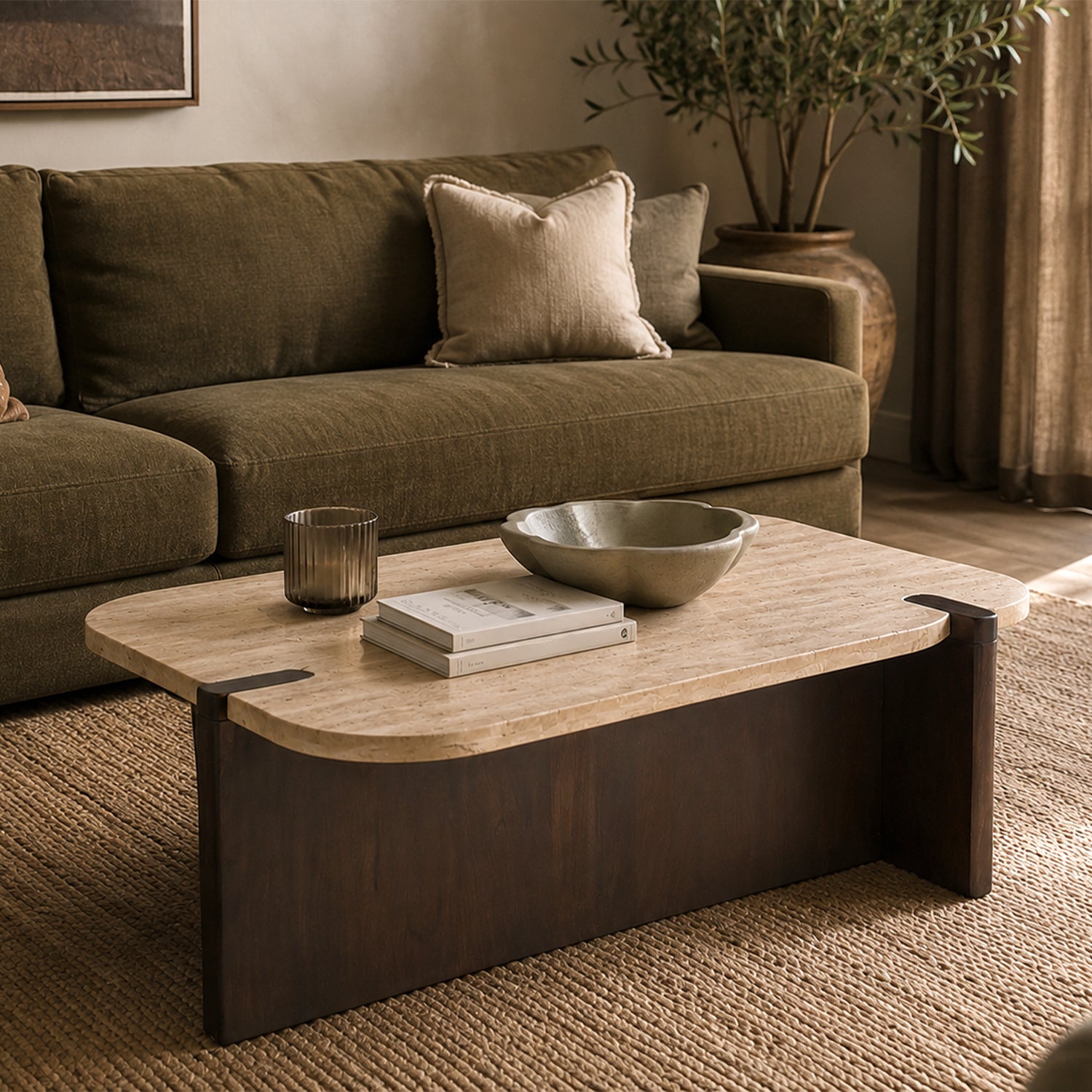 Carter Marble Coffee Table styled in front of an olive green sofa, featuring a natural travertine marble top with a sculptural dark timber base, accessorised with books, a decorative bowl, and a glass candle holder in a warm modern living room.