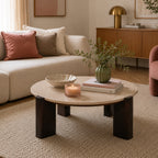 Round coffee table with a cream marble top and chunky dark brown timber legs, styled in a contemporary living room with decorative books, a green vase, a candle, and neutral furnishings.
