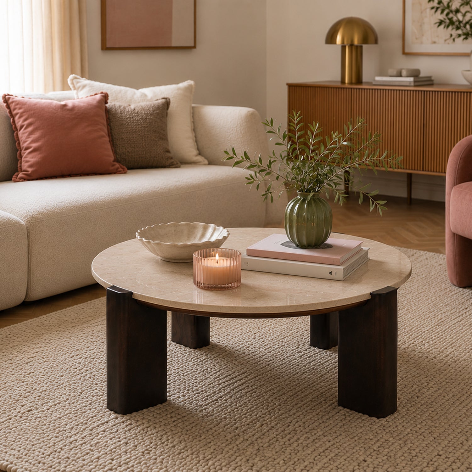 Round coffee table with a cream marble top and chunky dark brown timber legs, styled in a contemporary living room with decorative books, a green vase, a candle, and neutral furnishings.